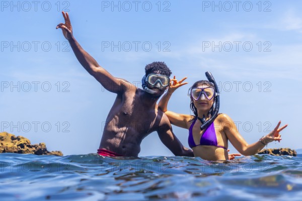 Young multi ethnic couple wearing snorkeling masks and enjoying summer vacation in tropical sea making victory hand gesture