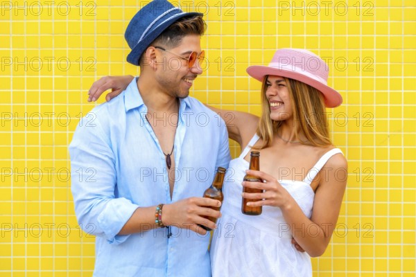 Young tourists drinking bottled beer, wearing summer clothes and hats, smiling and having fun on a hot summer day against a yellow tiled wall