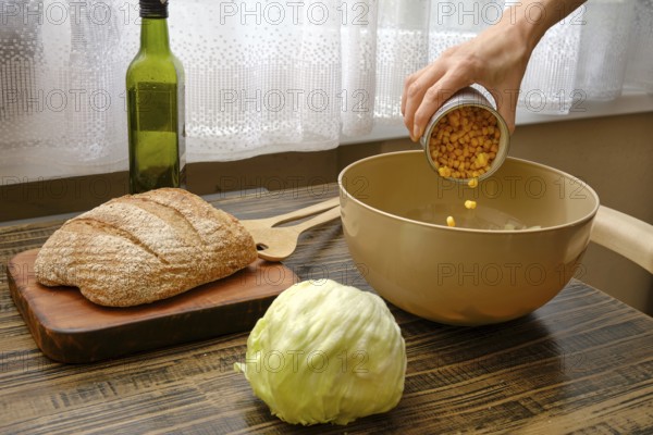 A hand pours canned corn into a mixing bowl on a wooden table. Nearby, a head of lettuce and a loaf of bread add to the fresh ingredients