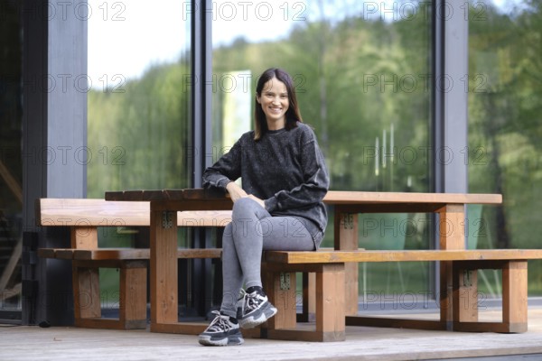 A woman with a bright smile is sitting at a rustic wooden bench on the porch of modern glass cabin, surrounded by greenery. She is wearing a cozy sweater and casual leggings