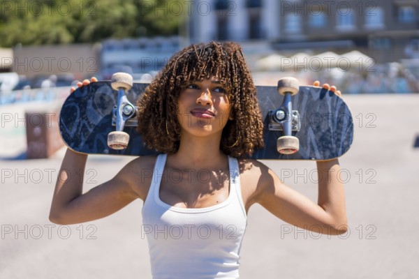 Confident young woman holding skateboard on shoulders, enjoying sunny day at skatepark, showcasing urban lifestyle and passion for skateboarding
