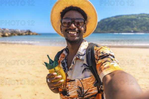 Young black man enjoying summer vacations on a tropical beach, taking a selfie while holding a refreshing pineapple cocktail