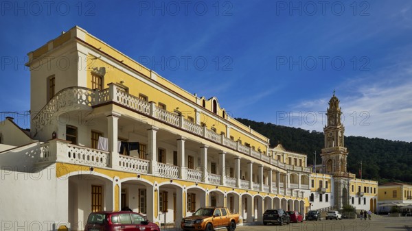 Yellow building with balconies, vehicles in front, blue sky in the background, Holy Week, Bay of Panormitis, Panormitis Monastery, Symi, Dodecanese, Greek Islands, Greece