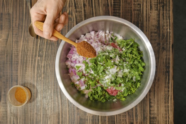 A hand holds a wooden spoon filled with spices over a stainless steel bowl containing chopped onions, herbs, and minced meat. The setting reflects a cozy kitchen atmosphere, emphasizing food preparation