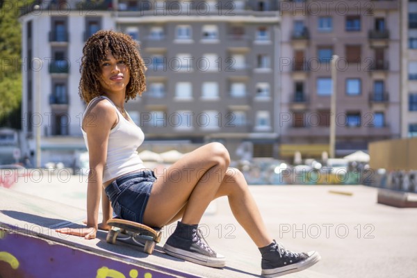Young woman sitting on her skateboard atop a ramp in an urban skatepark, soaking up the sun on a vibrant summer day