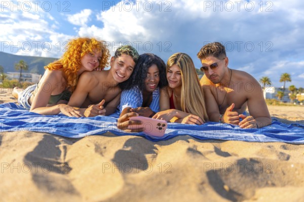 Five cheerful young friends are lying on a beach towel, taking a selfie with a smartphone, enjoying their summer vacation together