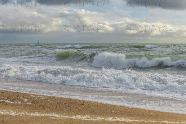 Powerful ocean waves wash over a sandy beach, creating white foam against a backdrop of dramatic clouds and distant land. The scene evokes a sense of calm and the strength of nature. Les Sables d'Olonne, Vendee, France