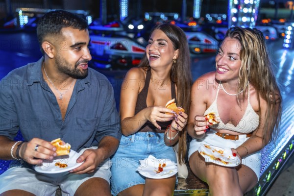 Three friends are enjoying hot dogs together at an amusement park, laughing and having a good time