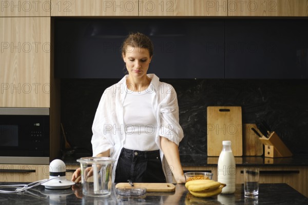 A woman stands in a contemporary kitchen, preparing to blend banana and sea buckthorn smoothies. Fresh ingredients are laid out on the counter, showcasing an inviting and healthy cooking activity