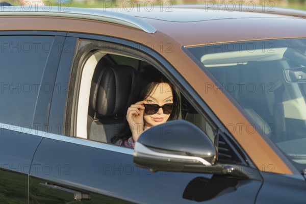 A woman with long dark hair wears stylish sunglasses while sitting in a sleek black car. The sun casts a warm glow, highlighting her features against a backdrop of greenery