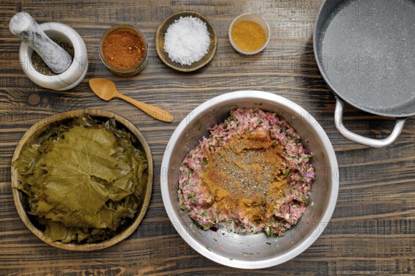 Preparing dolma involves mixing ground meat with spices and herbs on a rustic kitchen table. Fresh grape leaves and various seasonings are neatly arranged for rolling and cooking