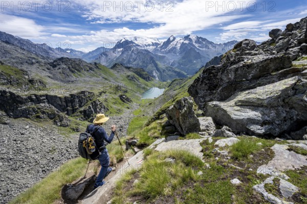 Mountaineer on hiking trail in mountain landscape, descent at Col du Louvie, view of mountain lake Lac du Louvie and glaciated mountain peaks of the Grand Combine, Valais Alps, Val de Bagnes, Valais, Switzerland