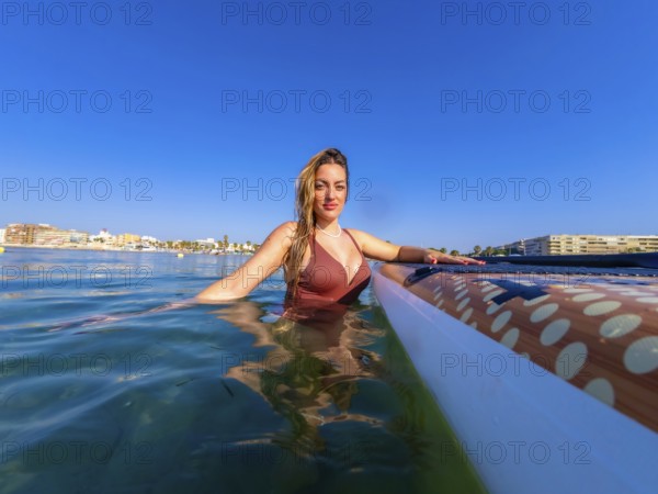 Woman wearing swimsuit relaxing in the sea next to her paddle board on a sunny summer day with a clear blue sky