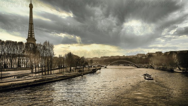 Under a dramatic sky, the Eiffel Tower stands tall beside the river Seine. Boats glide along the water while the evening light casts a beautiful glow on the Parisian landscape. Paris. France