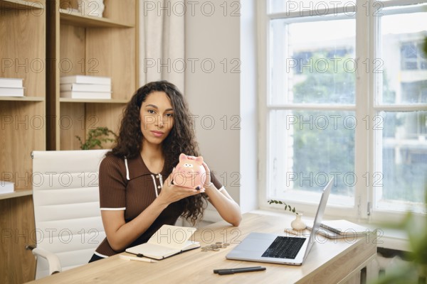 A woman with long curly hair sits at a desk in a bright home office, holding a piggy bank. She is surrounded by a laptop, notepad, and coins as she focuses on her financial planning