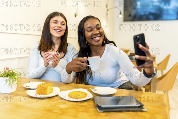 Two happy female friends are taking a selfie with smartphone while enjoying coffee and cake in a modern cafe