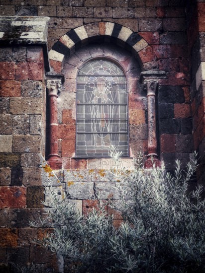 An ornate stained glass window featuring detailed artwork is set into a weathered stone wall. Surrounding vegetation adds a touch of nature to this historic architectural feature