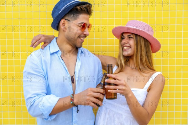 Young stylish tourists toasting with beer bottles, enjoying summer vacation together against a vibrant yellow tiled wall