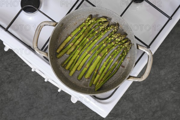 Fresh green asparagus is being cooked in a pan on a gas stove in the kitchen. The texture of the vegetables is highlighted