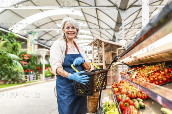 Senior greengrocer wearing gloves holding a shopping basket and smiling in a supermarket with fresh vegetables