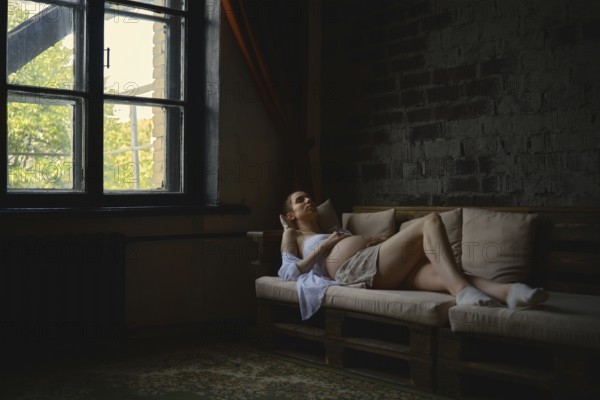A pregnant woman lounges comfortably on a cozy couch in her home. The warm sunlight filters through the window, creating a serene atmosphere