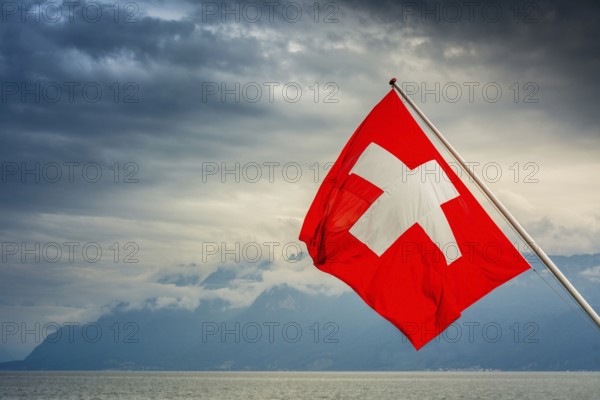 View of the Swiss flag waving over Lake Geneva with mountains and clouds in Switzerland during a cloudy day
