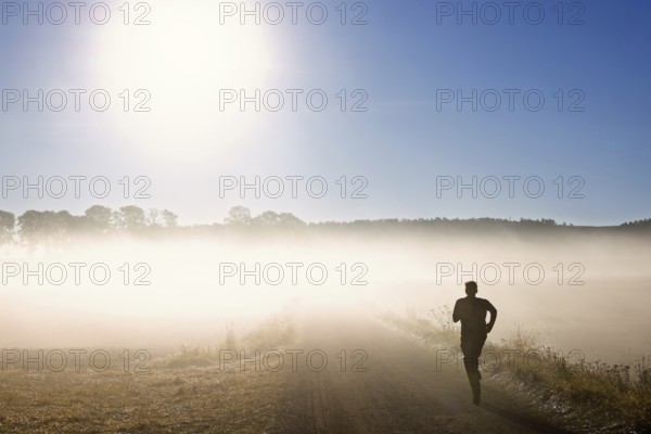Man training running on a gravel road in the morning fog in a rural landscape with the sun shining through the fog at autumn