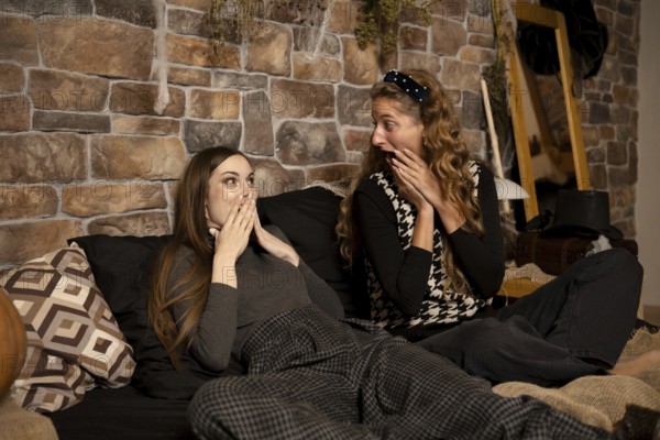 Two friends express surprise and excitement while lounging on a comfortable bed in a warmly decorated room. The ambiance creates a cozy atmosphere as they enjoy their time together