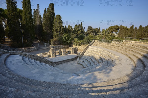 Ancient round amphitheatre with stone steps, surrounded by tall trees and ruins, Ancient Theatre, Odeon, Kos Town, Kos, Dodecanese, Greek Islands, Greece
