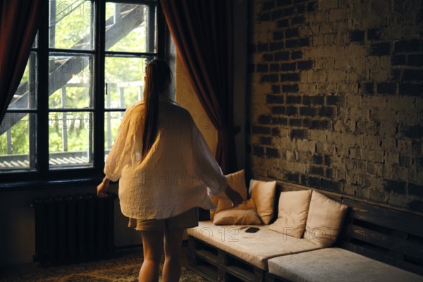 Rear view of a woman walking across the room to the sofa. The setting features exposed brick walls, wooden furniture, and a cozy atmosphere