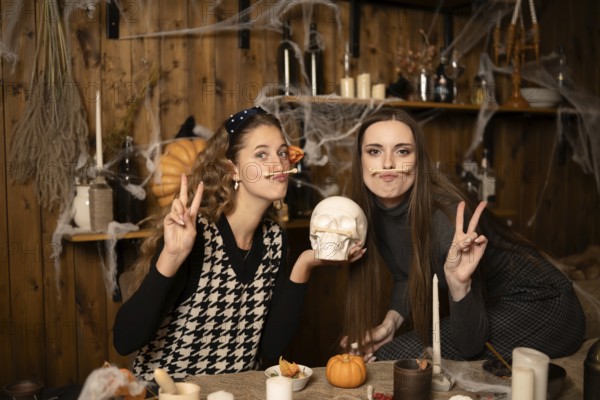 Two friends pose playfully in a decorated room for Halloween, surrounded by candles, pumpkins, and spider webs. They smile while holding props, creating a festive and fun atmosphere for the holiday