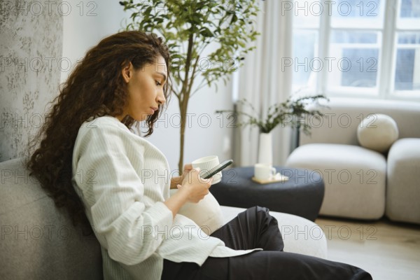 A woman sits on a comfortable sofa in a bright living room, sipping from a cup while focused on her smartphone. Natural light filters through large windows, enhancing the peaceful atmosphere
