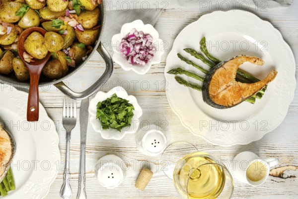 A beautifully arranged meal featuring a salmon fillet on asparagus, accompanied by seasoned potatoes with onions. Fresh herbs and a glass of white wine complete this inviting dining experience