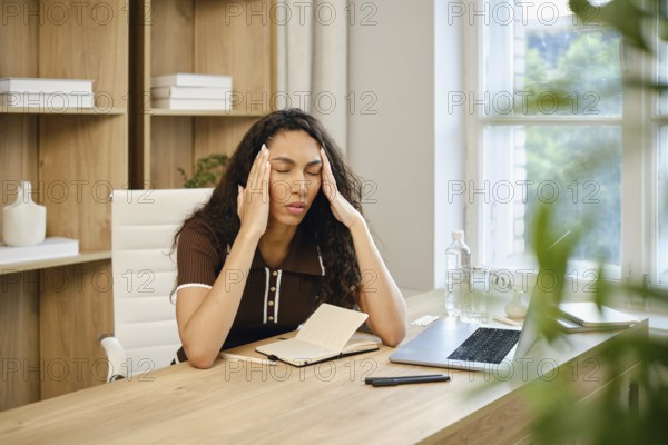 A woman with curly hair sits at a wooden desk in a stylish home office, visibly stressed while holding her head. She massages her temples with her fingers and closes her eyes