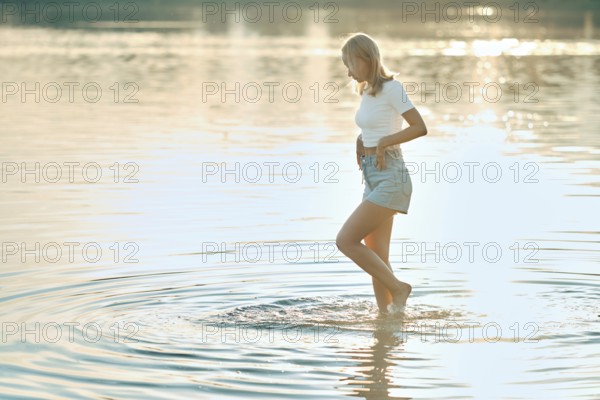 A woman is walking barefoot in gently rippling water at a tranquil lake during sunset. The warm light creates a peaceful atmosphere, highlighting her carefree spirit and connection to nature