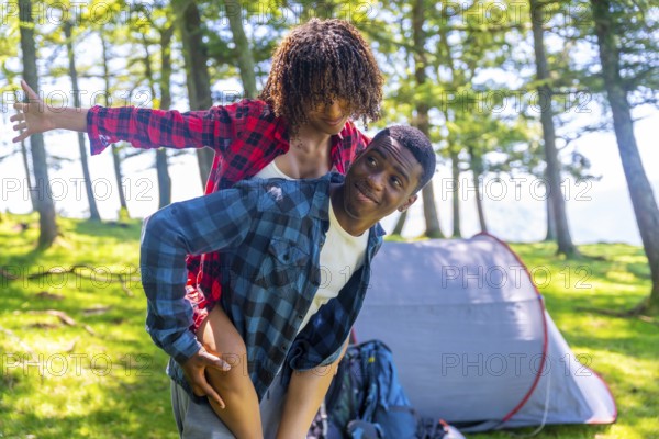 Young man giving piggyback ride to girlfriend in forest near tent, enjoying camping trip in nature