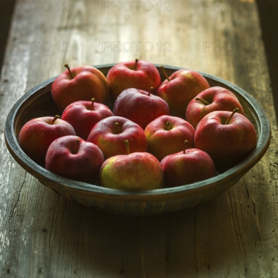 A rustic wooden bowl filled with freshly harvested red apples sits atop a weathered wooden table. The scene showcases the vibrant colors of the apples and the natural textures of wood