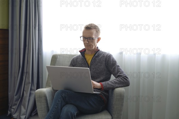 A man wearing glasses sits comfortably in a gray chair and fills out a tax return online on his laptop. Sunlight streams through the window, creating a bright and inviting workspace in the room