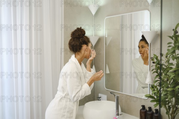 A woman in a white robe is applying skincare in a stylish bathroom. She is focused on her reflection in the mirror, creating a serene and personal moment. Plants add a touch of freshness