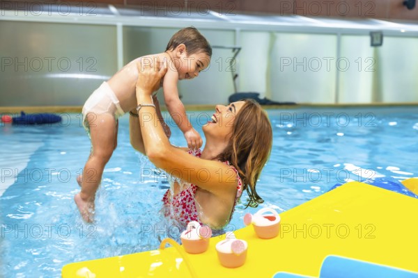Mother playfully lifting smiling baby out of water in indoor swimming pool during matinee swim lesson