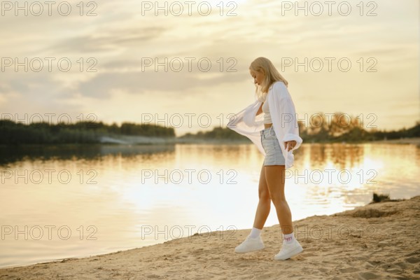 A young woman with long blonde hair walks along the sandy beach during sunset. She is happy to be on vacations