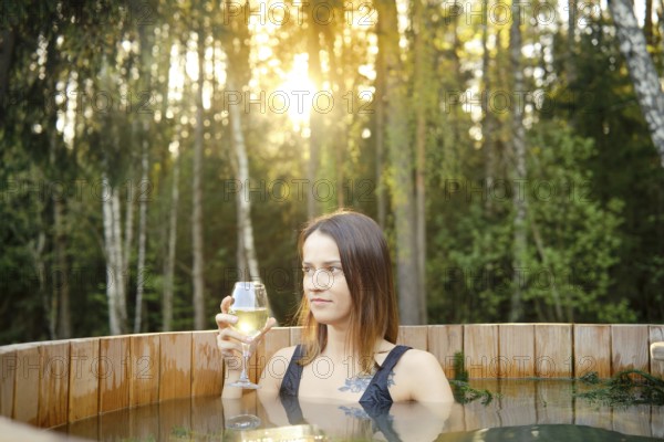 A woman is soaking in a wooden hot tub surrounded by tall trees, holding a glass of white wine. The sun sets in the background, casting a warm glow on the peaceful scene
