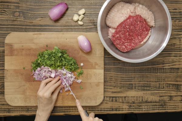A person chops fresh herbs and onions on a wooden cutting board while ground meat rests in a stainless steel bowl nearby