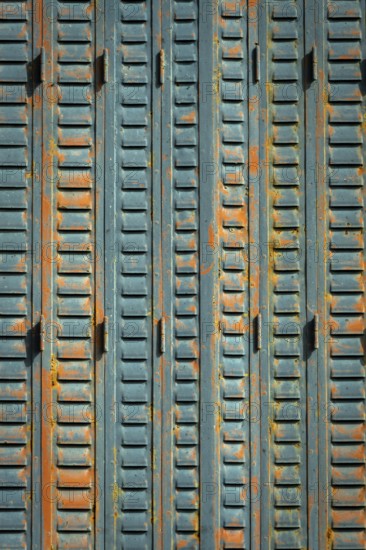 A close-up view of weathered metal shutters displaying a mix of blue paint and rust. The surface texture reveals signs of age, contributing to a rustic urban ambiance