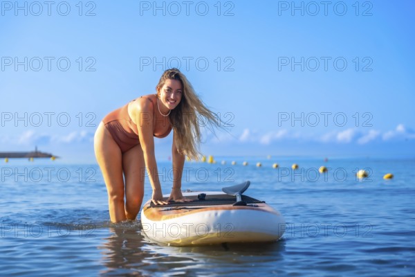 Smiling woman paddle surfing in calm blue waters, enjoying a sunny summer day. Clear sky and distant horizon create a peaceful backdrop