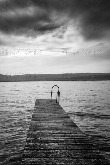 A rustic jetty stretches into the calm waters of Lake Geneva, framed by gentle waves and majestic mountains. The sky is adorned with soft clouds, creating a serene and tranquil atmosphere, Haute Savoie, Auvergne Rhone Alpes, France