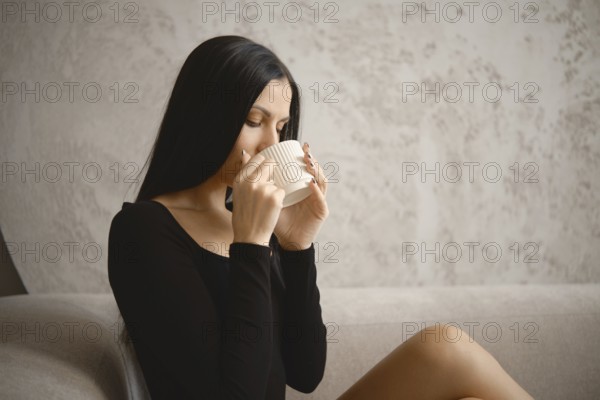 A young woman with long hair savors a warm drink, sitting comfortably on a stylish sofa in a contemporary living room. The soft lighting creates a serene atmosphere, perfect for relaxation