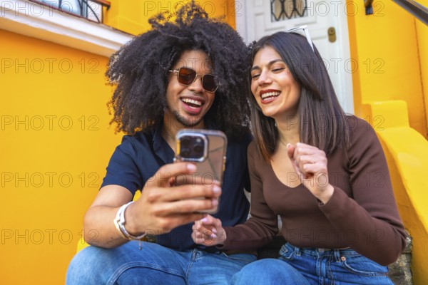 Two friends are sitting on the steps of a vibrant yellow building, laughing and watching a video on a smartphone