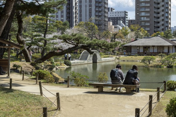 Tourists on a bench in Shukkei-en with blossoming cherry trees, Sakura, Seifukan teahouse and Koko-kyo stone bridge over Takuei-chi pond in spring, historical Japanese strolling garden, Japanese garden, green area, tourist attraction, sight, Naka-ku, Hiroshima, Japan