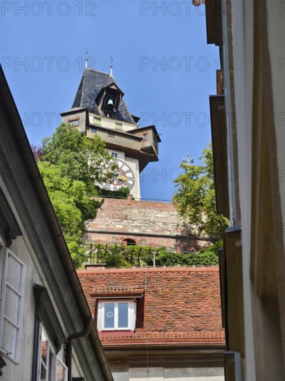 Historic clock tower towers over rooftops, surrounded by green trees and blue sky Graz Clock Tower Graz Styria Austria